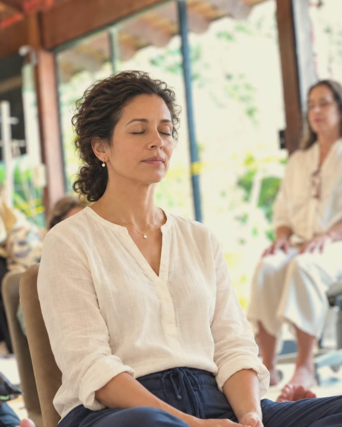 Uma mulher de olhos fechados, sentada em meditação na varanda da Lumin'área, com o grupo ao fundo